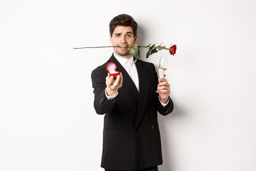 Romantic young man in suit making a proposal, holding rose in teeth and glass of champagne, showing engagement ring, asking to marry him, standing against white background.