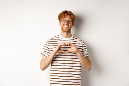 Valentines day. Happy boyfriend with red hair, smiling and showing heart gesture, I love you, standing over white background.
