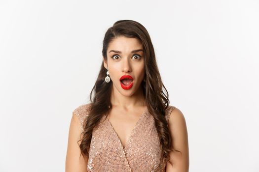 Image of attractive woman with red lips and hairstyle, wearing dress, staring surprised at camera, standing over white background.