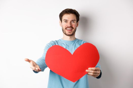 Handsome man in love making confession to you, pointing hand at camera, holding big red heart cutout on valentines day, singing romantic serenade, standing over white background.