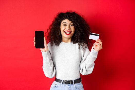 E-commerce and online shopping concept. Cheerful woman smiling, showing plastic credit card and empty smartphone screen, standing on red background.