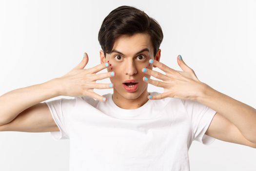 People, lgbtq and beauty concept. Close-up of beautiful gay man with glitter on face, showing nail polish on his fingernails, looking sassy at camera, standing over white background.