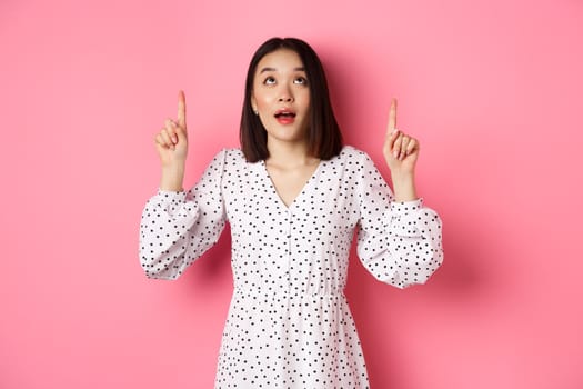 Image of beautiful asian girl in dress looking, pointing fingers up, gazing dreamy at promo offer, standing over pink background.