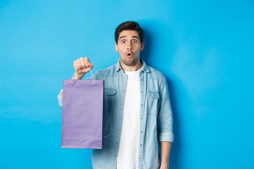 Concept of shopping, holidays and lifestyle. Handsome surprised guy holding paper bag from shop and looking amazed, standing over blue background.