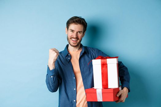 Cheerful young man got discounts on valentines day, making fist pump and say yes, holding gift boxes with presents for lover, standing over blue background.