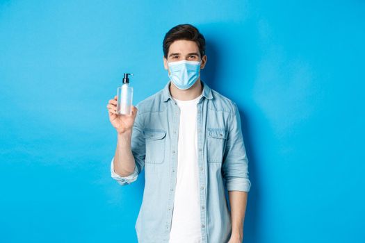 Concept of covid-19, pandemic and social distancing. Young man in medical mask showing hand sanitizer, standing against blue background.
