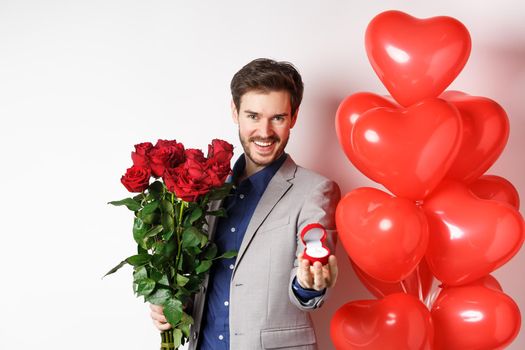 Handsome boyfriend in suit making a wedding proposal, showing engagement ring and say marry me, holding red roses, standing near Valentines day balloons, white background.
