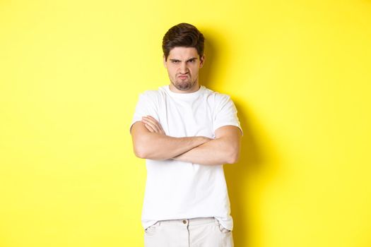 Offended and mad guy, frowning and sulking, holding hands crossed on chest, standing angry against yellow background.