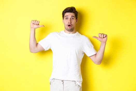 Surprised handsome guy pointing at himself, looking amazed, standing over yellow background. Copy space