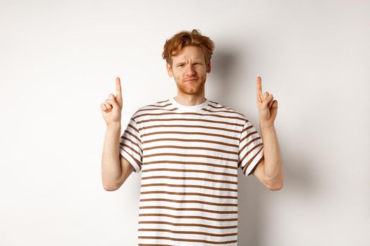 Skeptical handsome redhead man pointing fingers up, grimacing and looking doubtful at camera, standing indecisive over white background.