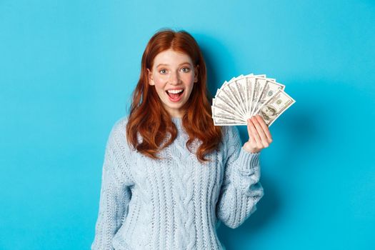 Happy redhead girl in sweater, staring excited at camera, showing money dollars, standing against blue background.