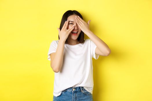 Intrigued asian girl waiting for surprise, peeking through fingers on eyes, smiling happy, standing over yellow background.