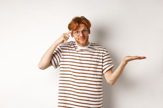 Are you stupid. Confused redhead guy in glasses shrugging, pointing at head and staring skeptical at camera, white background.