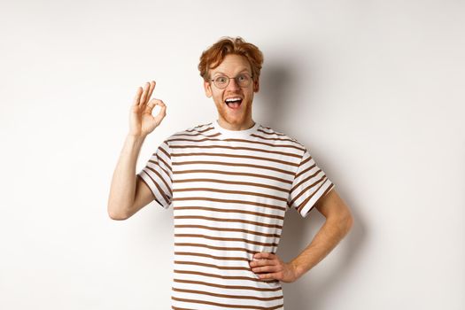 Excellent job. Happy redhead man praising you, showing OK sign and smiling amazed, approve and like something good, standing over white background.