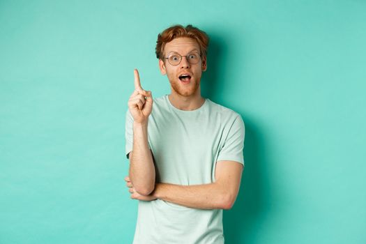 Excited young man with ginger hair in glasses, raising index finger, pitching an idea, standing over mint background. Copy space