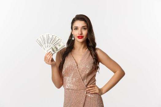 Shopping concept. Sassy beautiful woman with red lipstick, wearing glamour dress, showing money dollars and looking confident, standing over white background.