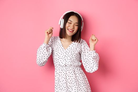 Happy young asian woman dancing and having fun, listening music in headphones, standing over pink background. Copy space