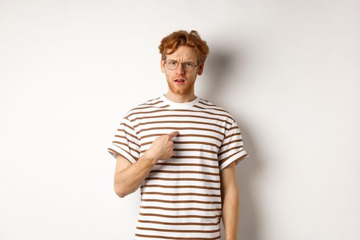 Confused redhead man in glasses pointing at himself, looking questioned at camera, white background.