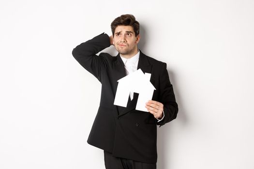 Image of indecisive businessman thinking, holding house maket and looking at upper left corner doubtful, standing against white background.