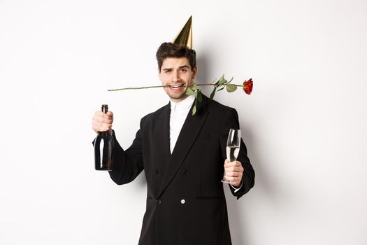 Romantic guy in trendy suit, celebrating and having a party, holding rose in teeth and champagne, standing over white background.