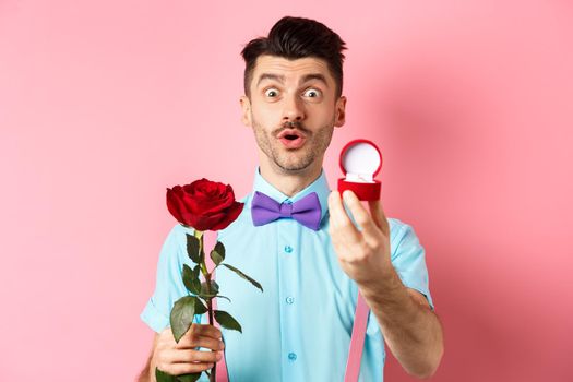 Valentines day. Funny man with moustache and bow-tie making proposal, showing engagement ring and propose with red rose, standing over pink background.