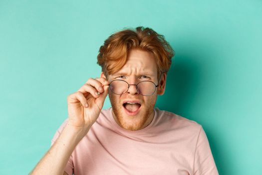 Close up portrait of redhead guy take-off glasses and looking confused at something strange, standing shocked over turquoise background.