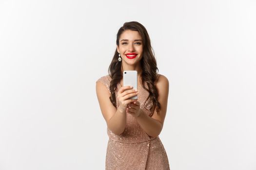 Beautiful young woman in party dress and makeup, taking photo on smartphone, shooting you on phone camera, standing over white background.