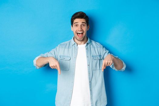 Image of excited handsome man pointing fingers down, making an announcement, standing against blue background.