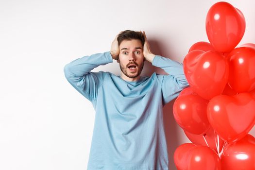 Confused boyfriend holding hands on head and panicking on Valentines holiday, alarmed with romantic presents on lovers day, standing near hearts balloon over white background.