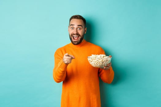 Handsome young man holding bowl of popcorn, smiling amazed and pointing at camera, standing over blue background.