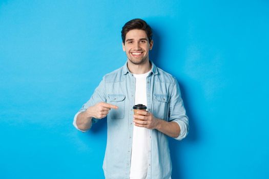 Smiling happy man pointing at paper cup with coffee, recommending cafe, standing over blue background.