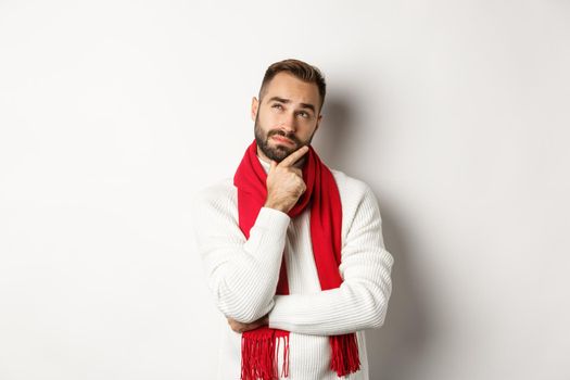 Christmas holidays and celebration concept. Thoughtful young man with beard thinking, looking at upper left corner, making choice of gift, standing over white background.