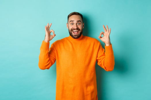 Impressed man praising something awesome, showing okay signs and looking amazed, standing in orange sweater at light blue background.