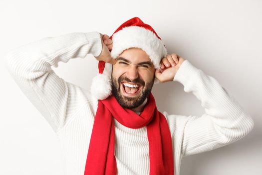 New Year party and winter holidays concept. Close-up of cheerful bearded man celebrating Christmas, smiling and wearing santa hat, white background.
