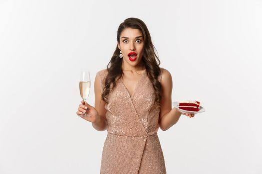 Party and celebration concept. Elegant woman with red lips, glamour dress, drinking champagne and eating cake, staring surprised at camera.