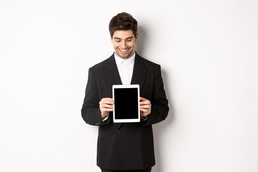 Image of good-looking male entrepreneur in black suit, looking down at digital tablet screen and showing advertisement, standing against white background.