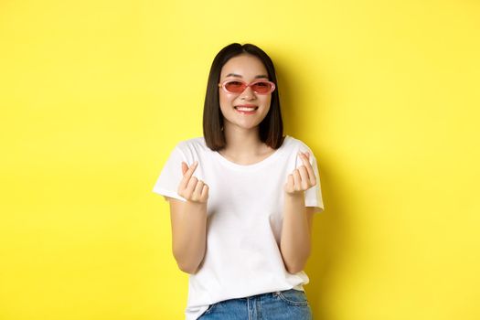 Fashion and lifestyle concept. Attractive asian woman in stylish sunglasses, showing finger hearts and smiling happy at camera, standing over yellow background.