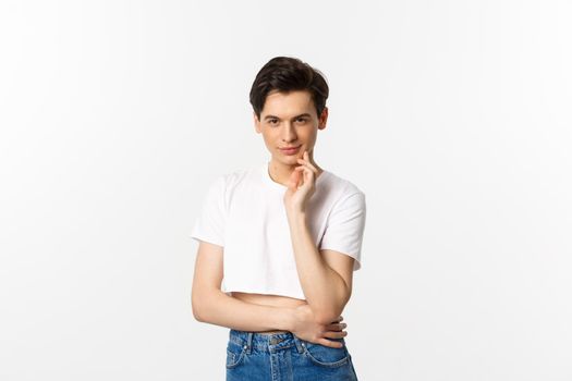 Thoughtful young queer man wearing crop top, smiling and looking at camera cunning, having an idea, standing over white background.