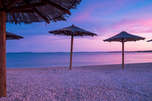 Beach umbrellas, view of the beautiful blue and purple sunset, sky and straw beach umbrellas. Perfect holiday concept. Travel and vacation. Beach on Vir Island, Croatia, Europa. Copy space.