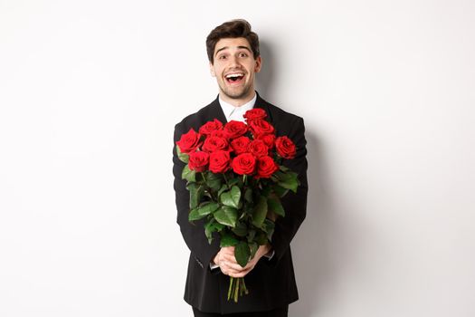 Image of handsome boyfriend in black suit, holding bouquet of red roses and smiling, being on a date, standing over white background.