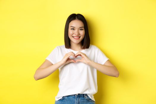 Beautiful asian woman showing I love you heart gesture, smiling at camera, standing against yellow background. Concept of valentines day and romance.
