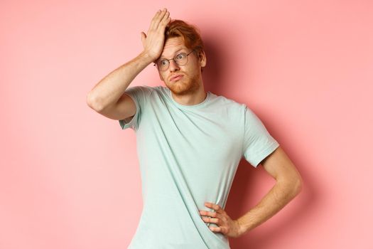 Tired young man with red hair, wearing glasses, looking annoyed and tensed, making facepalm gesture and exhale bothered, standing over pink background.