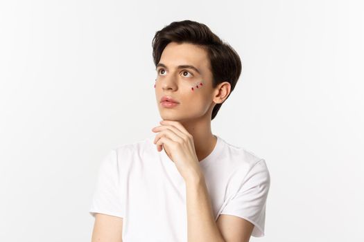 People, lgbtq community and lifestyle concept. Beautiful androgynous man with glitter on face, looking at upper left corner sensual, standing over white background.