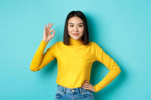 Beautiful smiling asian woman recommend product, showing Ok sign and looking satisfied, standing over blue background.