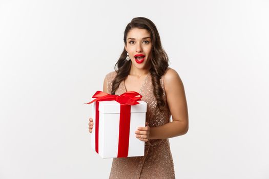 Merry Christmas. Image of beautiful woman in glamour dress receiving gift and looking surprised, celebrating New Year, standing over white background.