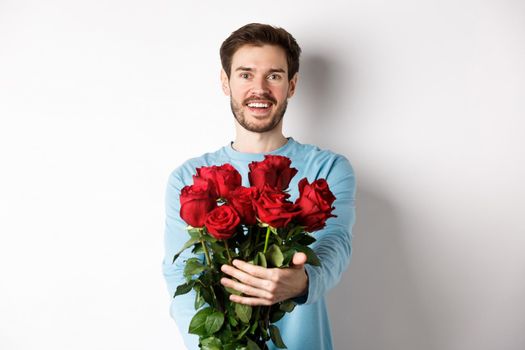 Handsome bearded guy stretch out hands, giving bouquet of roses and smiling, bring flowers on romantic date, celebrating Valentines day with lover, standing over white background.