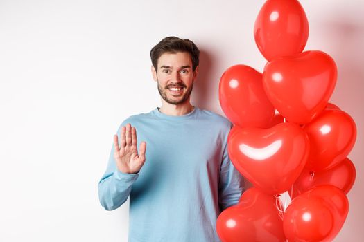 Handsome boyfriend saying hello, bring romantic red heart balloons on date, waving hand and smiling, standing over white background.