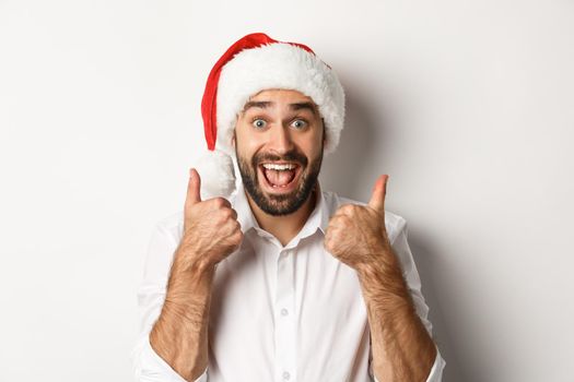 Party, winter holidays and celebration concept. Man enjoying christmas, wearing santa hat and showing thumb up with excited face, white background.