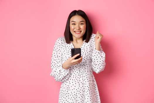 Online shopping and beauty concept. Excited asian woman winning in internet, holding smartphone and rejoicing, smiling happy and celebrating, standing over pink background.