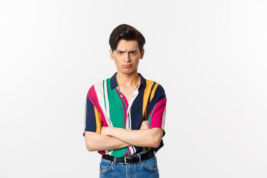 Offended and angry young gay man sulking, cross arms chest and looking judgemental at camera, standing over white background.